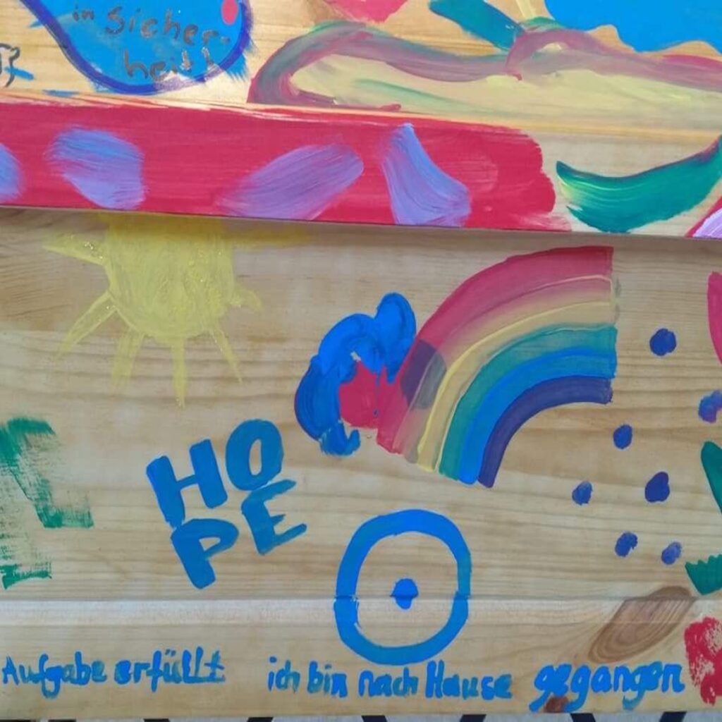 Nahaufnahme eines handbemalten Sarges. Der helle Kiefernsarg ist mit bunten Farben, Sonne und Regenbogen bemalt. Er ist beschriftet mit dem Wort Hope und dem Satz: Aufgabe erfüllt. Ich bin nach Hause gegangen.