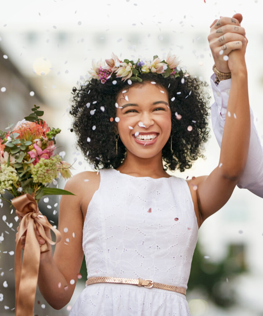 Eine Braut im weißen Kleid mit Lochstickerei und schmalem, goldenen Gürtel hält einen Blumenstrauß mit Schleife und strahlt über das ganze Gesicht. Sie hat dunkle, krause Haare mit Blumen geschmückt und eine hellbraune Haut. Um sie herum fliegt Konfetti und sie sieht sehr glücklich aus.