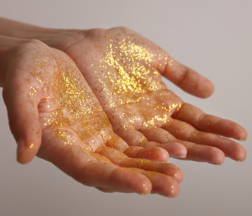 Vor einem hellen Hintergrund sind zwei empfangende Hände sichtbar. Sie haben eine helle Hautfarbe und sind mit goldenem Glitzerstaub bedeckt.
